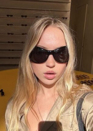 Long blonde hair styled with black oversized sunglasses and a tan jacket